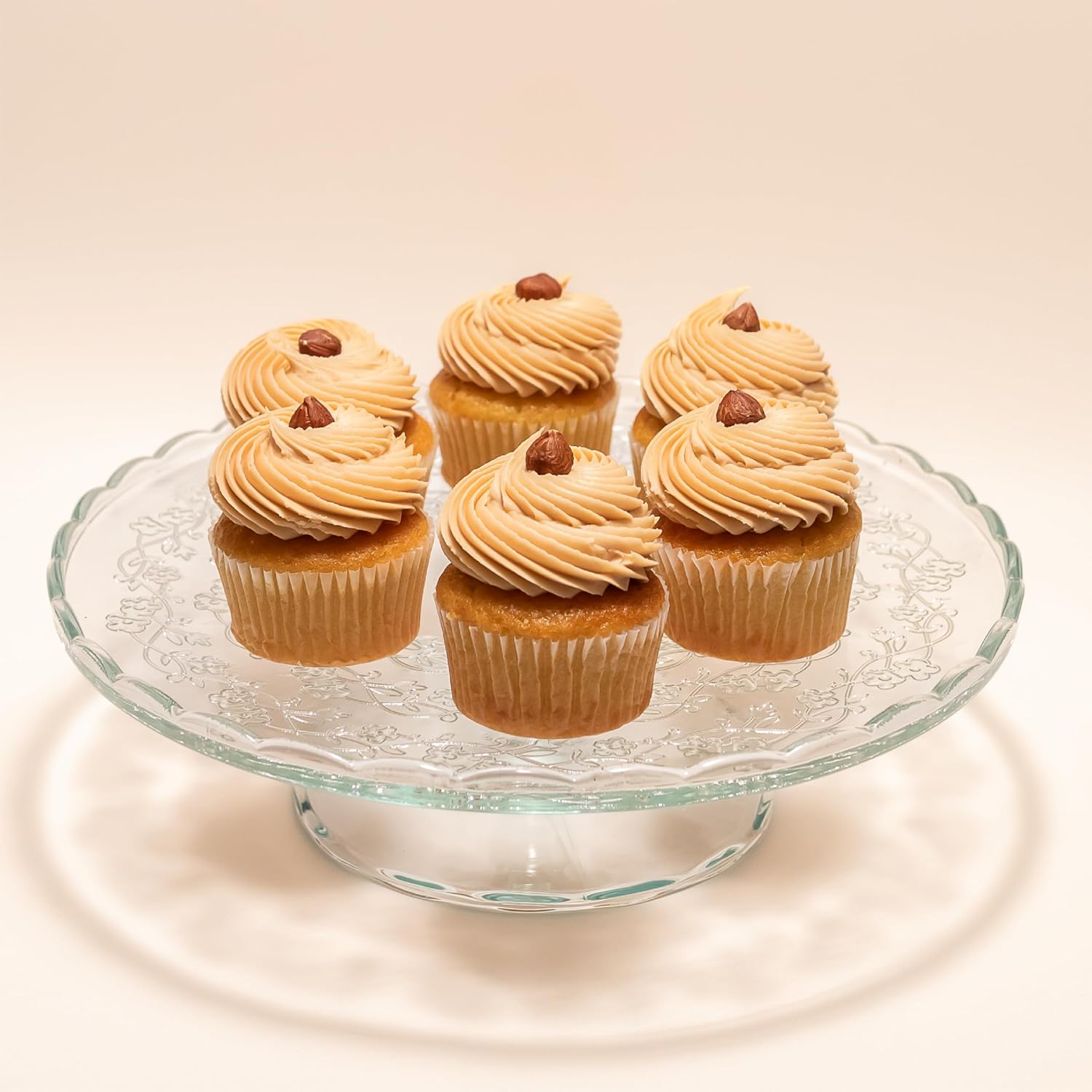 Six cupcakes displayed on a glass cake stand, each topped with a swirl of hazelnut cream frosting and garnished with a whole hazelnut.