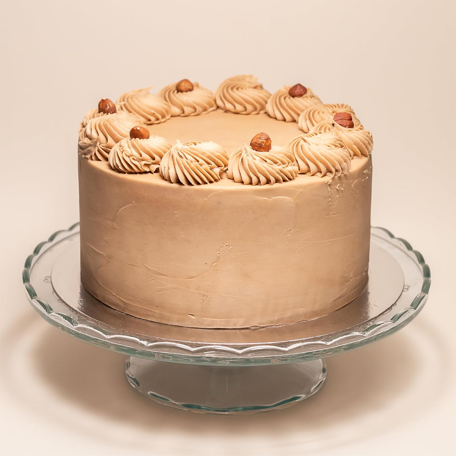 Whole cake covered in hazelnut cream frosting, decorated with swirls of frosting and whole hazelnuts around the top edge, displayed on a glass cake stand.