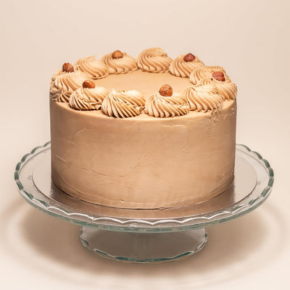 Whole cake covered in hazelnut cream frosting, decorated with swirls of frosting and whole hazelnuts around the top edge, displayed on a glass cake stand.
