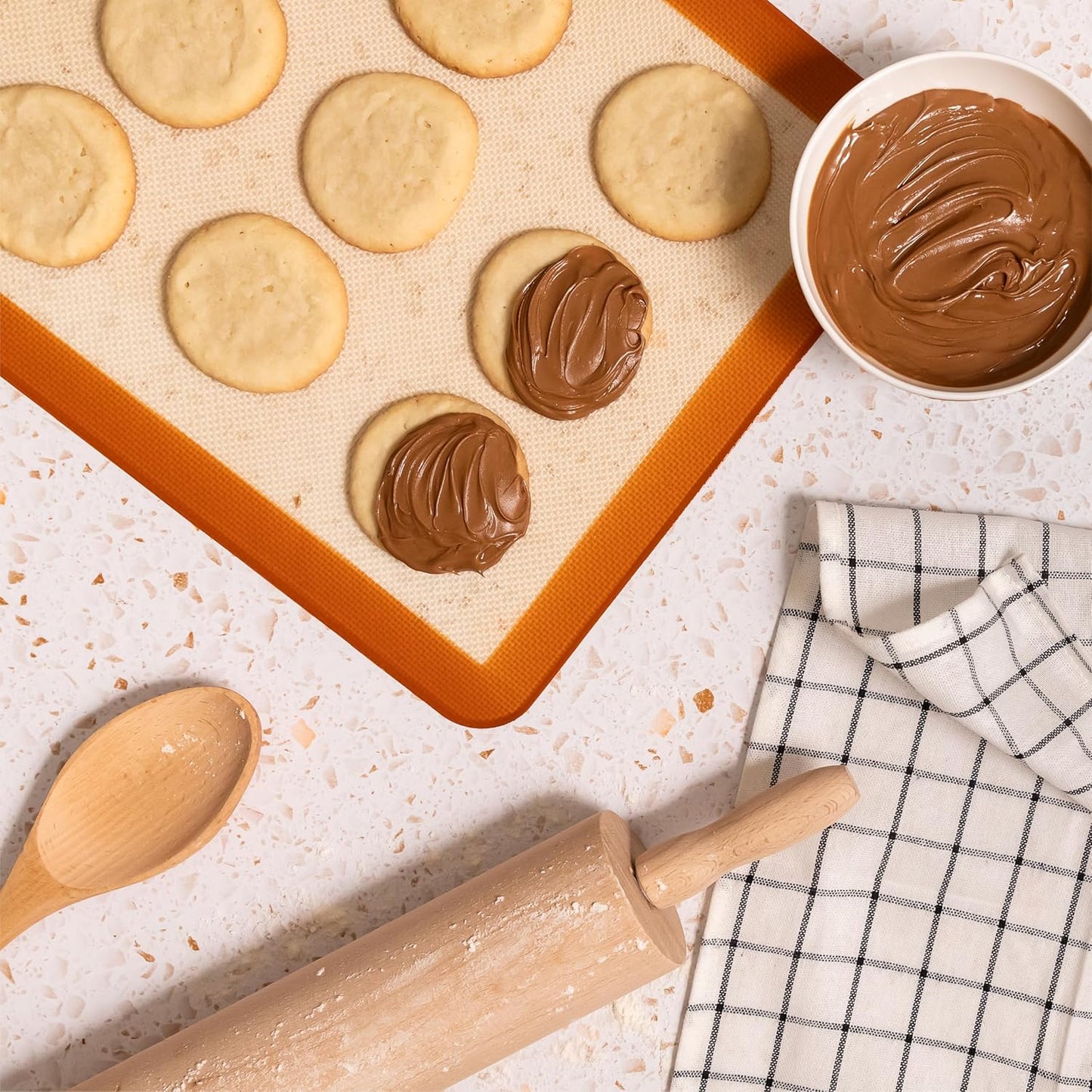 Freshly baked cookies on a baking mat, some topped with creamy hazelnut spread, with a bowl of spread, a rolling pin, wooden spoon, and checkered kitchen towel on the counter.
