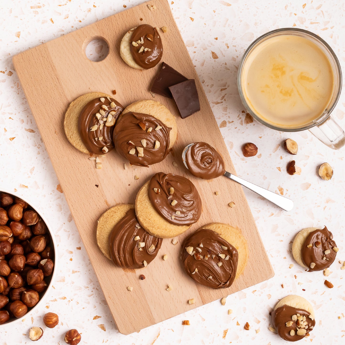 Cookies topped with creamy hazelnut spread and chopped nuts on a wooden board, served with a cup of coffee, chocolate pieces, and whole hazelnuts.