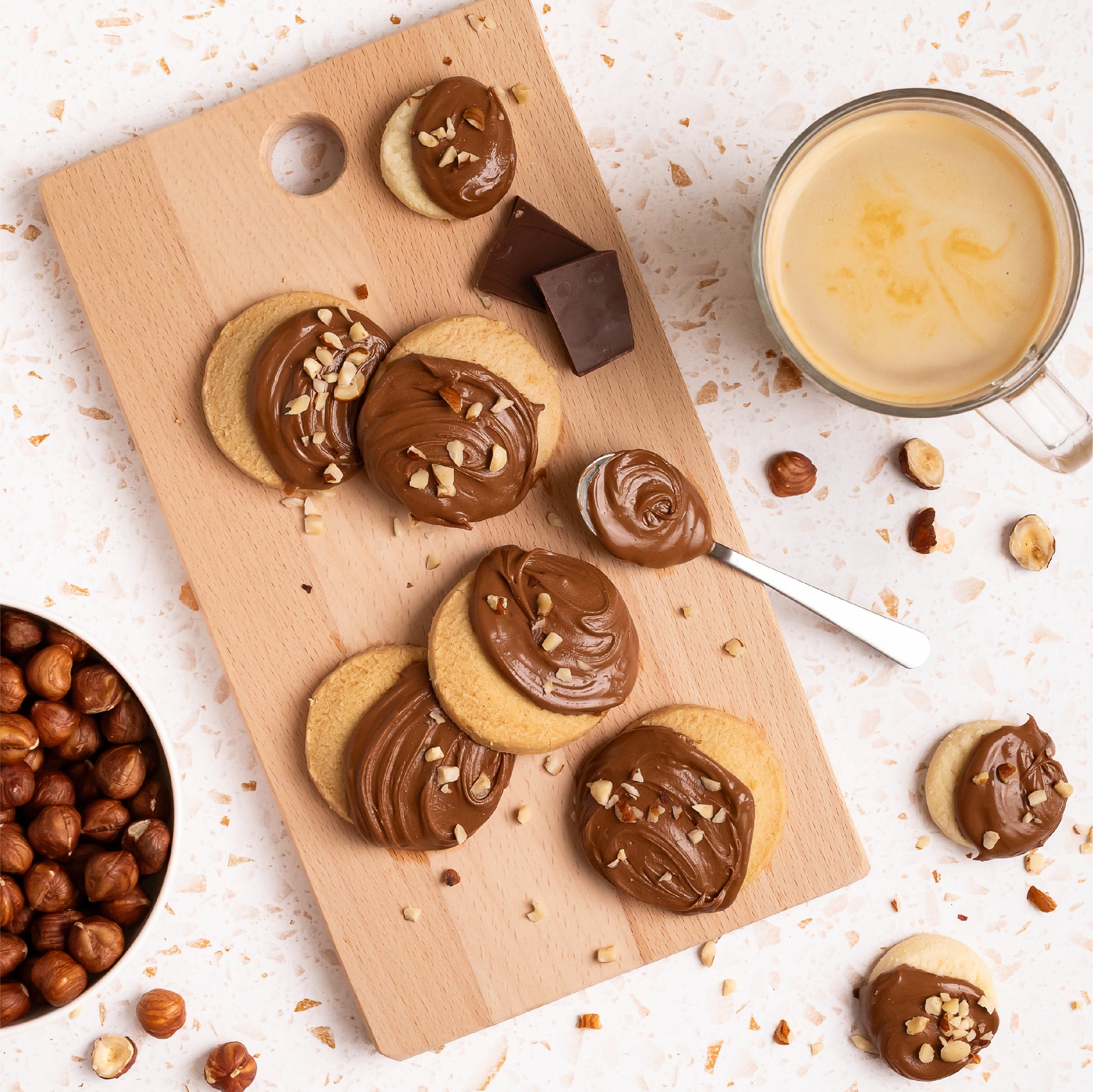 Cookies topped with creamy hazelnut spread and chopped nuts on a wooden board, served with a cup of coffee, chocolate pieces, and whole hazelnuts.