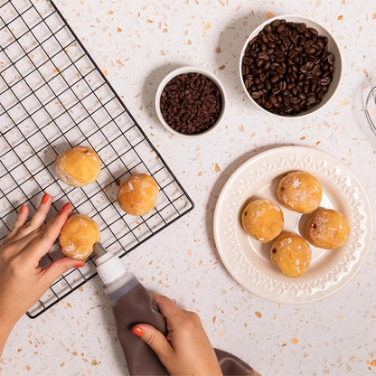 Mini donuts being filled with Nutlicious Hazelnut Mocha Spread using a piping bag, surrounded by bowls of chocolate chips and coffee beans on a light surface