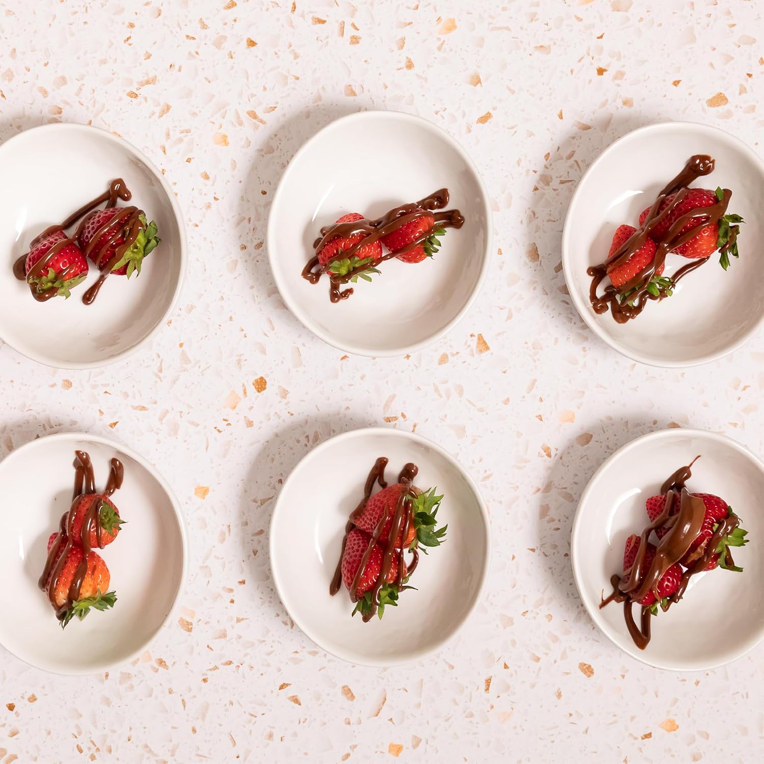 Six small white bowls with fresh strawberries drizzled with Nutlicious Hazelnut Mocha Spread on a terrazzo surface.