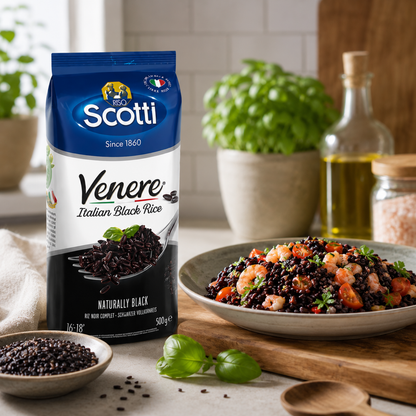 A bag of Scotti Venere Italian Black Rice is shown standing on a kitchen counter beside a prepared bowl of black rice salad with pasta and vegetables, with basil leaves, a bottle of olive oil, and other kitchen items in the background.