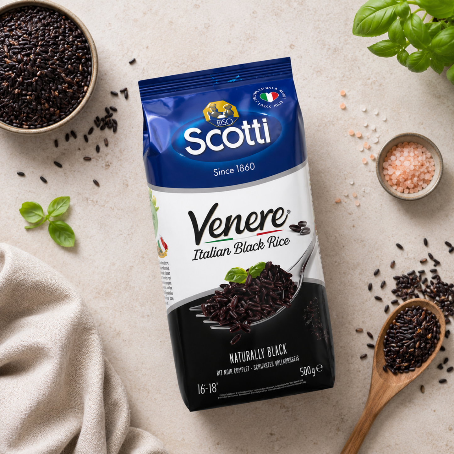 A 500g package of Scotti Venere Italian Black Rice is displayed in a styled flat-lay on a light beige surface, surrounded by ingredients like fresh basil, a wooden spoon of black rice, and bowls of salt and loose black rice.