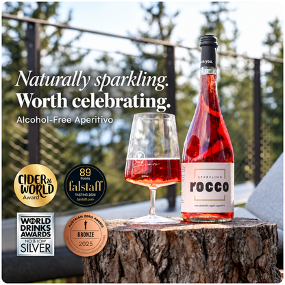 A bottle of Rocco Alcohol-Free Sparkling Aperitivo and a filled wine glass sit on a tree stump outdoors, with promotional text and award badges on the left side of the image.