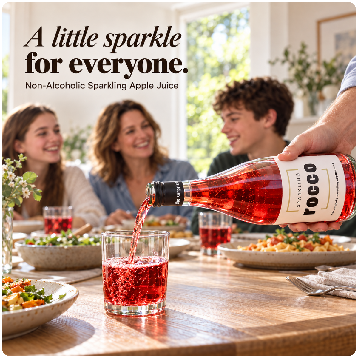 A bottle of Rocco sparkling apple juice is being poured into a glass on a dining table, with a family blurred in the background. Text at the top reads, 'A little sparkle for everyone.
