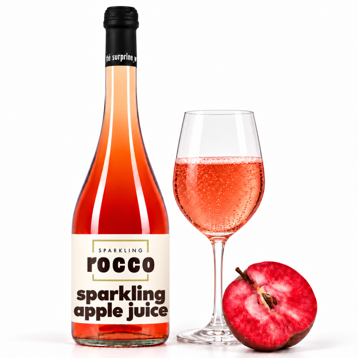 A bottle of Rocco Sparkling Apple Juice next to a filled wine glass and a halved red apple on a white background.