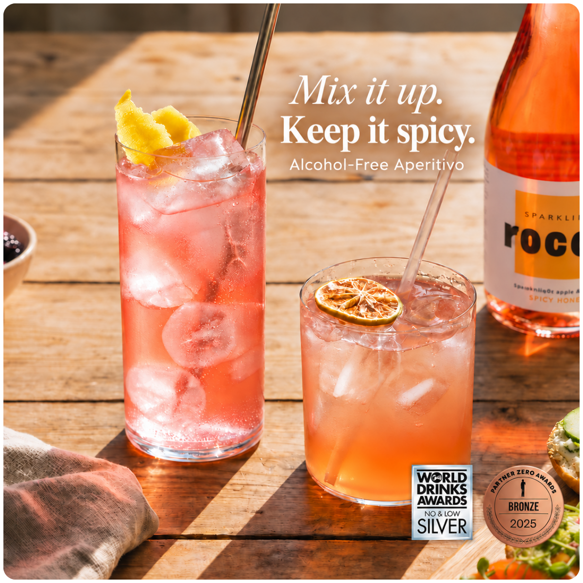 Two glasses of pink cocktail-style iced drink on a wooden table, with a bottle of sparkling rosé wine partially visible and promotional text reading 'Mix it up. Keep it spicy.
