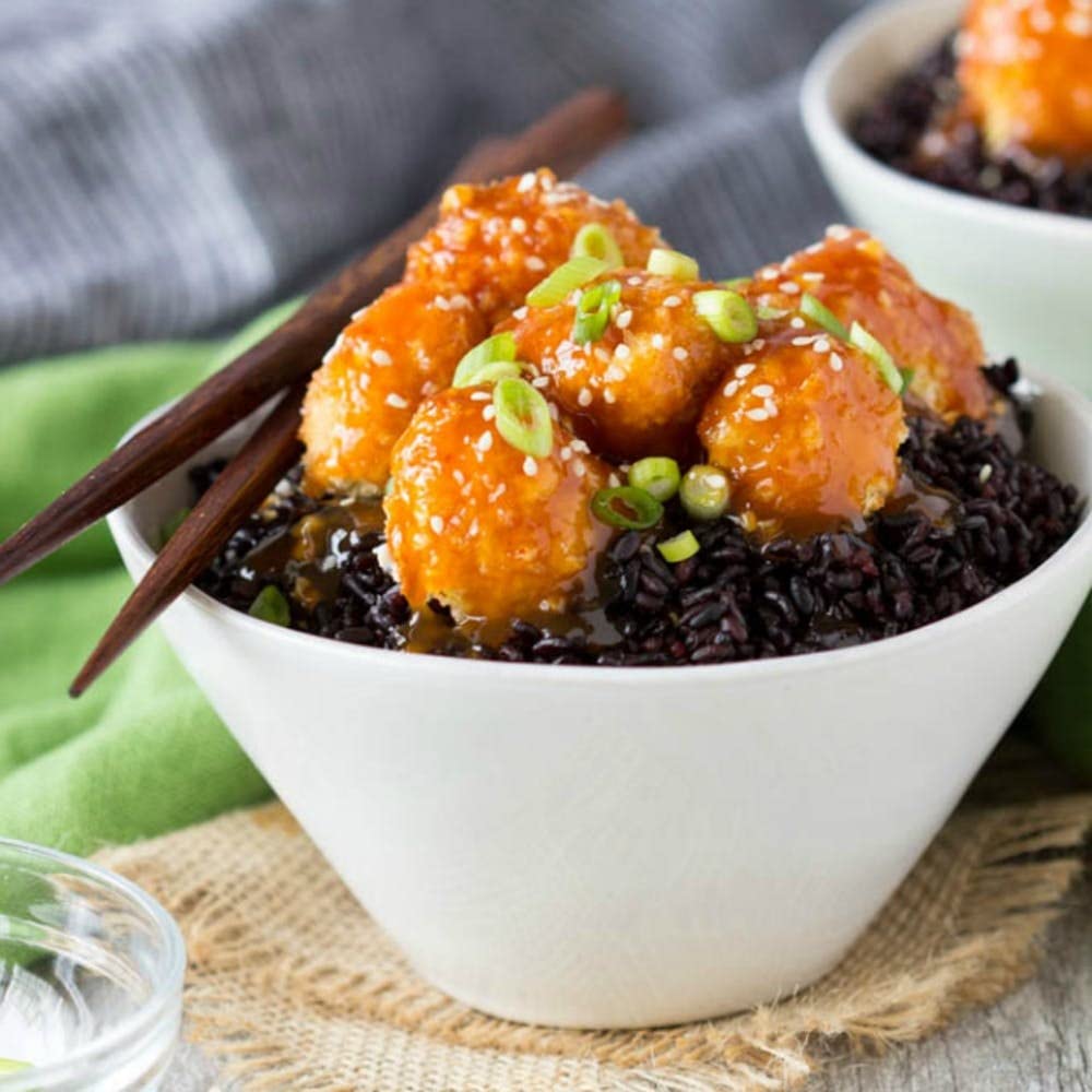 White bowl filled with Riso Scotti Venere wholegrain black rice, topped with crispy orange-glazed chicken bites, sesame seeds, and sliced green onions, with chopsticks resting on the bow