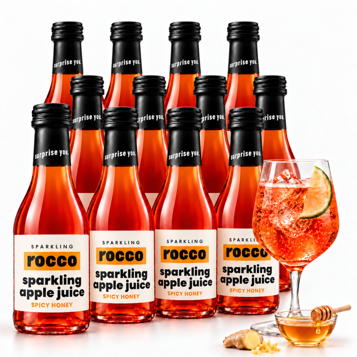 A group of Rocco Sparkling Apple Juice bottles with a glass of prepared orange-red drink with a lime wedge and honey dipper on a clean white background.