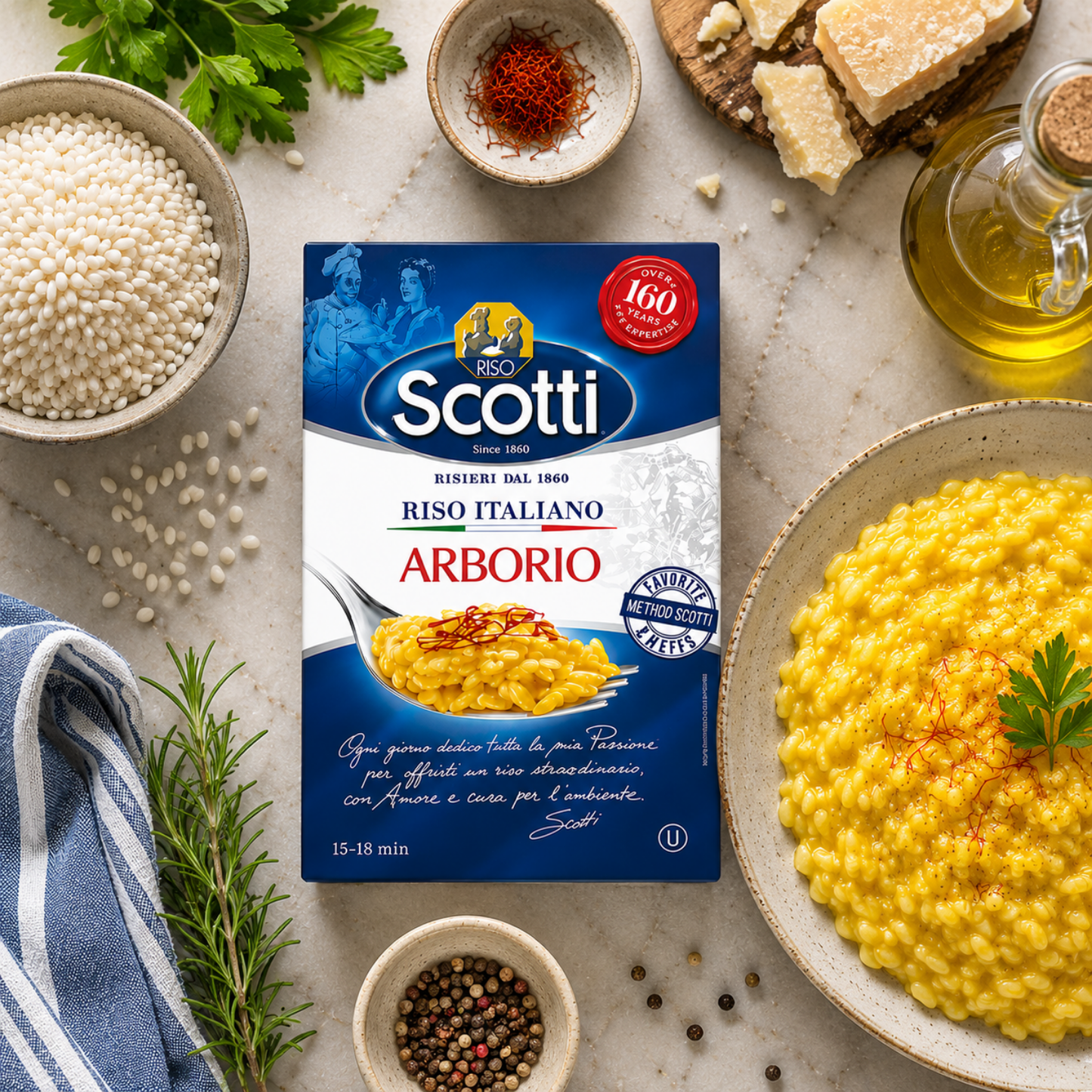 A package of Scotti Riso Italiano Arborio pasta is centered on a styled kitchen table surrounded by ingredients and prepared food. The package is blue and white with red accents, and the scene includes bowls of yellow risotto, rice grains, olive oil, herbs, and spices.
