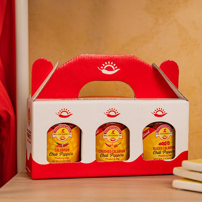 Red and white packaging box with three jars of TuttoCalabria calabrian chili peppers on a wooden surface.