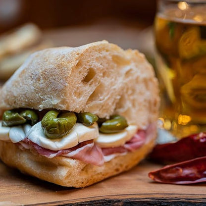 Rustic Italian sandwich with green olives, mozzarella cheese, and prosciutto on crusty bread, served on a wooden board with a drink in the background.