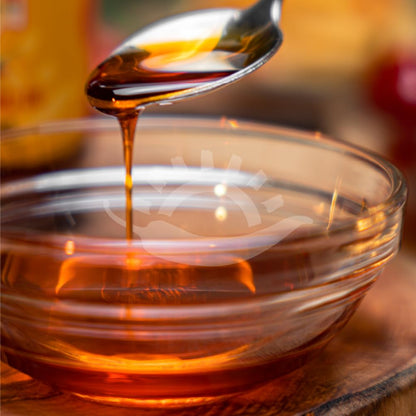 A close-up of amber honey dripping from a metal spoon into a clear glass bowl on a wooden surface, with a warm blurred background.