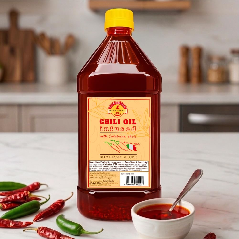 A bottle of Chili Oil Infused with Calabrian Chili by Golden Spice Company, placed on a kitchen counter with whole red and green chilies and a small bowl of the chili oil with a spoon.