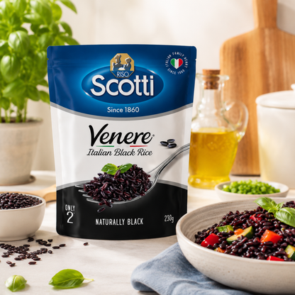 A bag of Scotti Venere Italian Black Rice stands on a kitchen counter with a bowl of cooked black rice, a small dish of green beans, a bottle of olive oil, and fresh basil leaves in a bright kitchen setting.