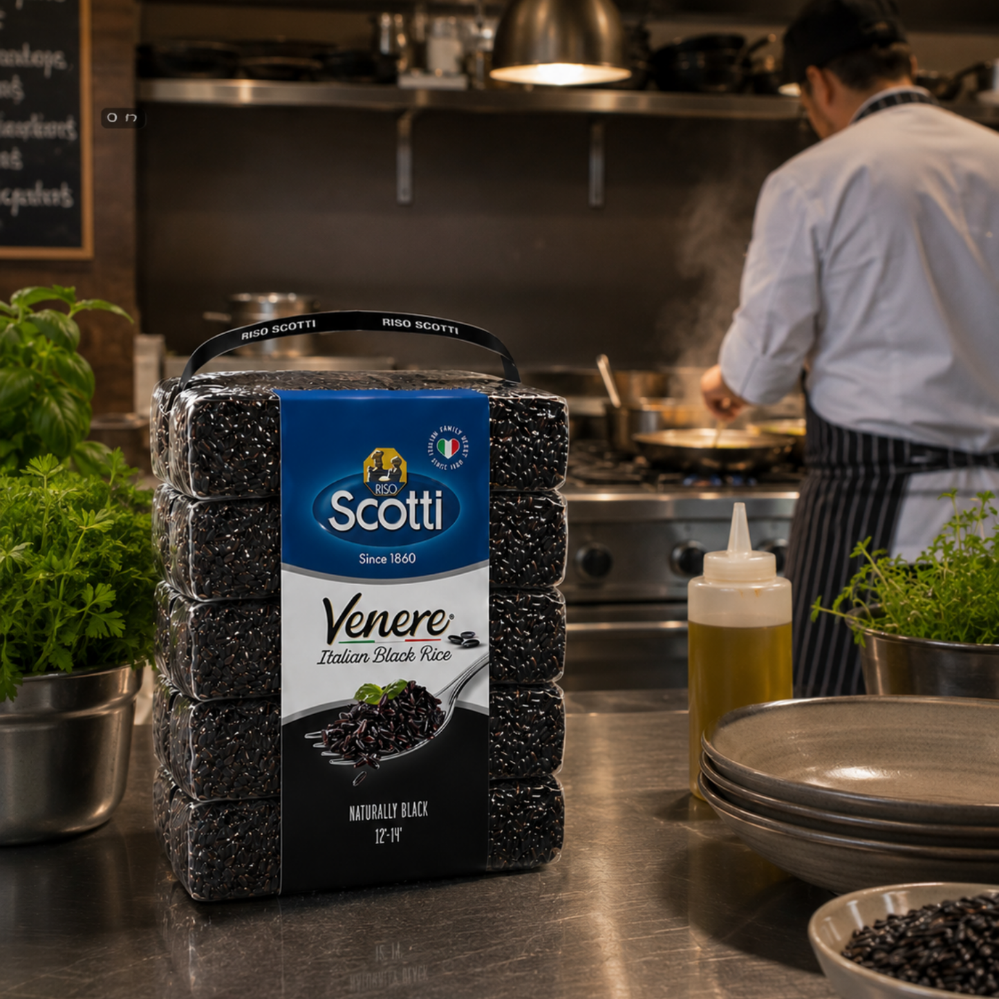 A package of Scotti Venere Morbido Black Rice (Italian Black Rice) sits on a stainless steel kitchen counter in a professional kitchen, with a chef cooking in the background.