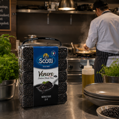 A package of Scotti Venere Morbido Black Rice (Italian Black Rice) sits on a stainless steel kitchen counter in a professional kitchen, with a chef cooking in the background.