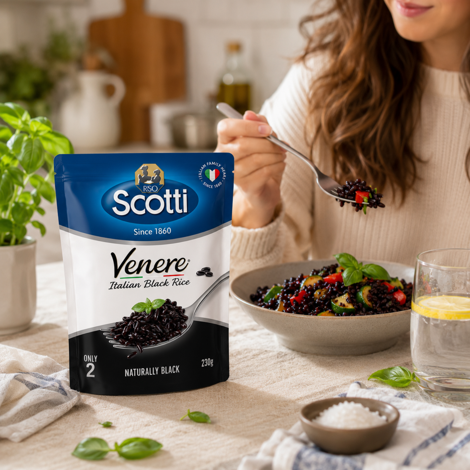 A bag of Scotti Venere Italian Black Rice stands on a kitchen table beside a bowl of prepared pasta, with a woman holding a spoonful of the black rice in the background.