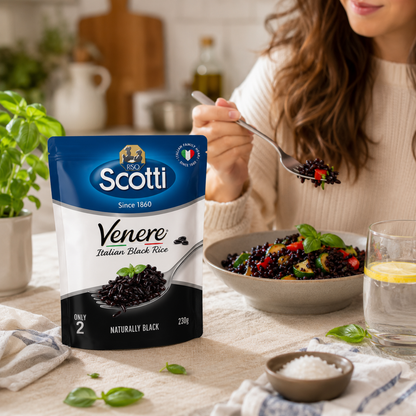 A bag of Scotti Venere Italian Black Rice stands on a kitchen table beside a bowl of prepared pasta, with a woman holding a spoonful of the black rice in the background.