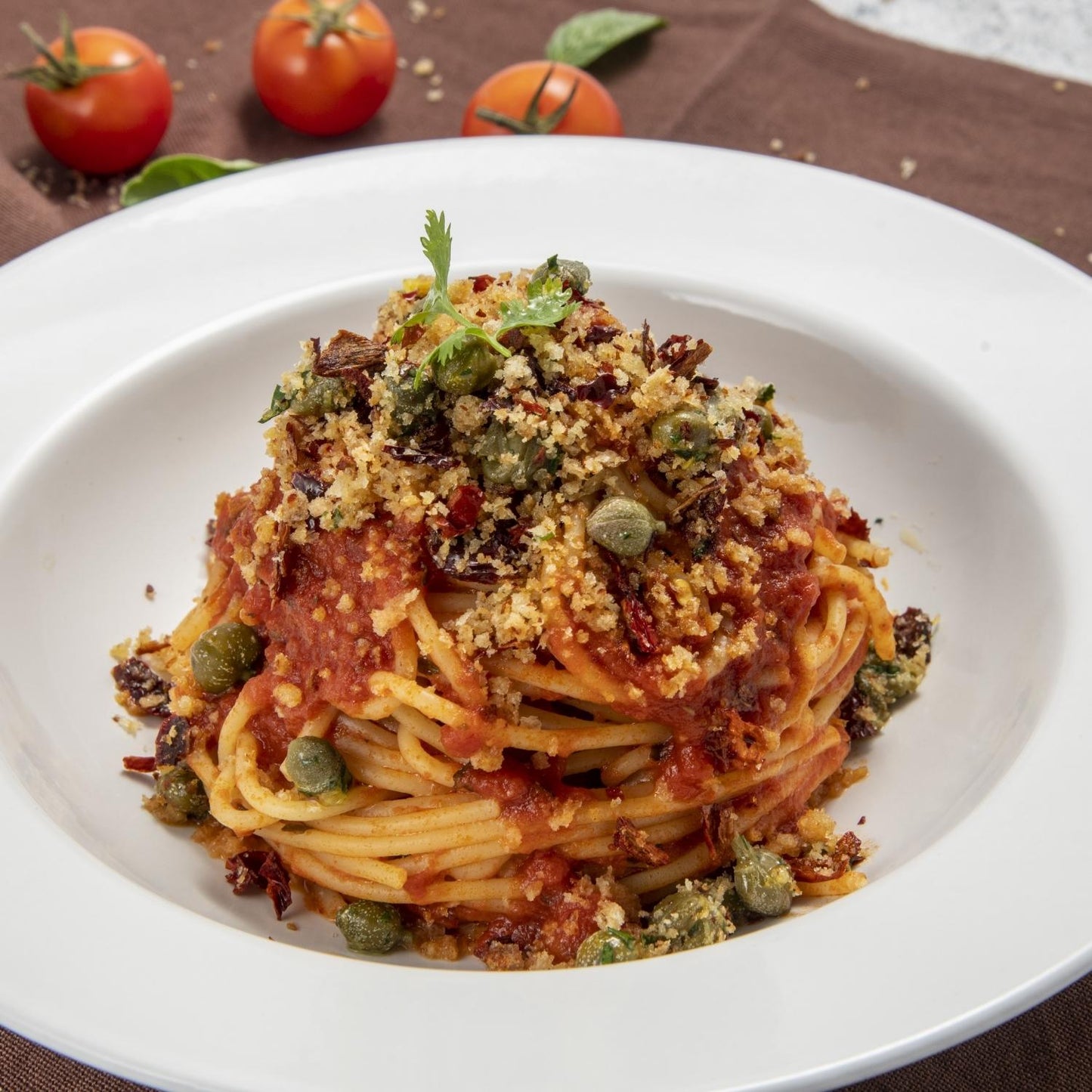 Plate of Italian spaghetti topped with tomato sauce, Fratelli D'Amico salted capers, breadcrumbs, and herbs, garnished with fresh tomatoes in the background.