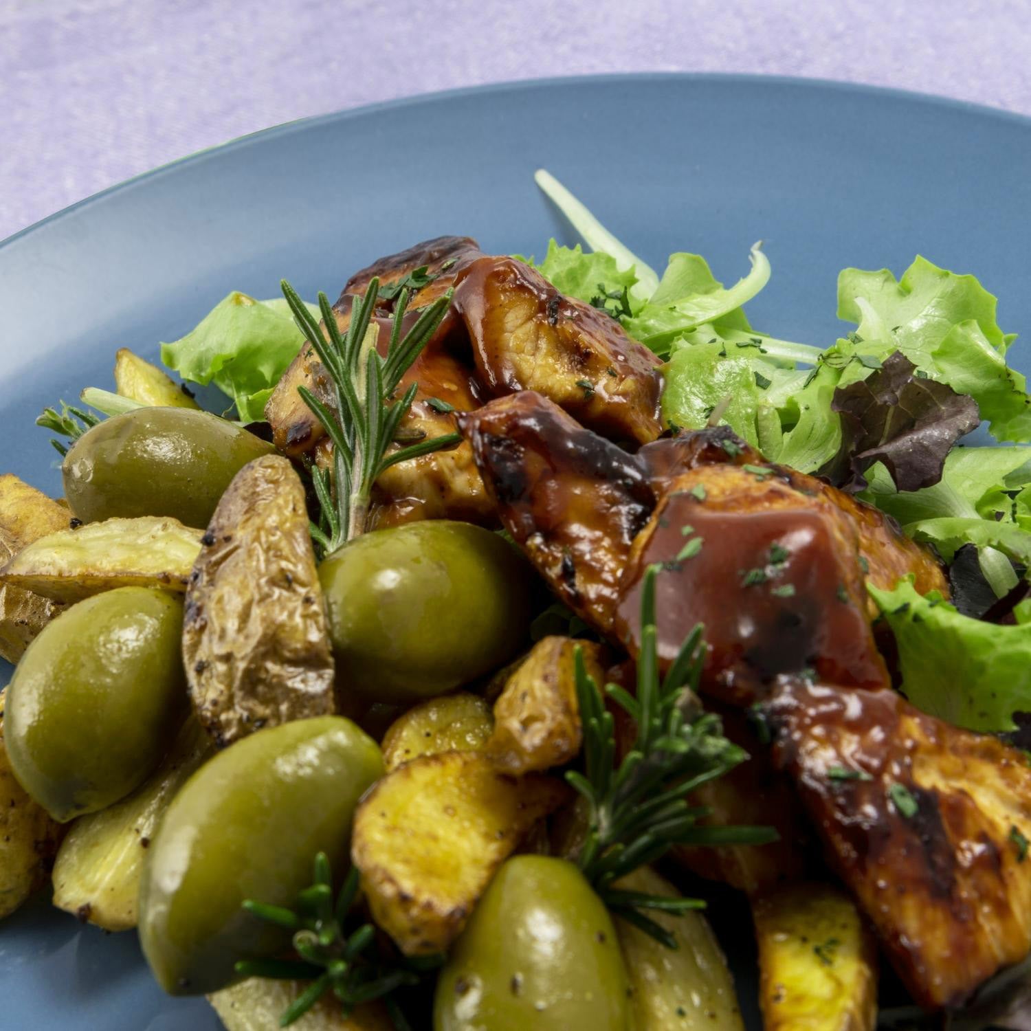 Close-up of a gourmet dish featuring roasted potatoes, large green Cerignola olives, grilled meat glazed with sauce, fresh salad greens, and garnished with rosemary on a blue plate