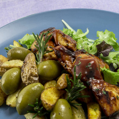 Close-up of a gourmet dish featuring roasted potatoes, large green Cerignola olives, grilled meat glazed with sauce, fresh salad greens, and garnished with rosemary on a blue plate