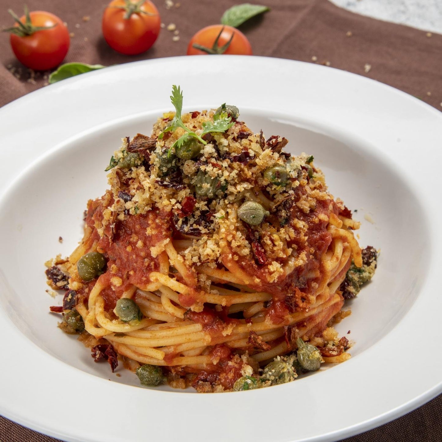 Plate of Italian spaghetti topped with tomato sauce, Fratelli D'Amico salted capers, breadcrumbs, and herbs, garnished with fresh tomatoes in the background.